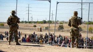 Migrants wait in line adjacent to the border fence under the watch of the Texas National Guard